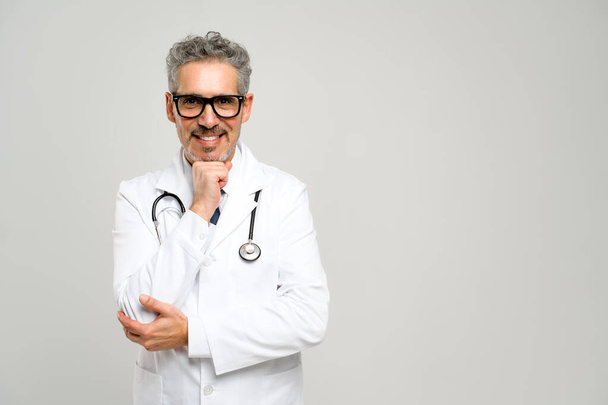 A senior grey-haired doctor stands confidently with a stethoscope around his neck, smiling warmly at the camera, exuding a sense of trust and professionalism. - Fotoğraf, Görsel