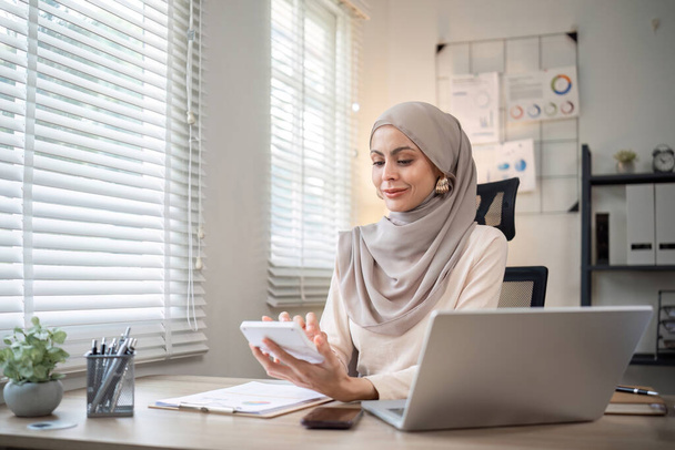 Ελκυστική μουσουλμάνα λογίστρια επιχειρηματίας που εργάζεται με τη χρήση φορητού υπολογιστή στο σύγχρονο γραφείο. - Φωτογραφία, εικόνα