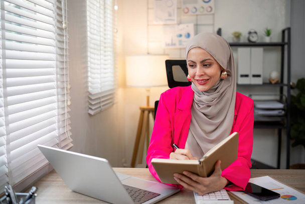 Geschäftsfrau asiatische Muslimin mit Taschenrechner und Schreiben Notizen mit berechnen Finanzen auf einem Schreibtisch im Büro. Frau arbeitet im Büro mit Laptop und Steuer, Buchhaltung, Unterlagen auf dem Schreibtisch. - Foto, Bild