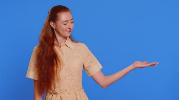 Redhead γυναίκα δείχνει αντίχειρες και δείχνει δεξιά κενό μέρος, διαφημιστική περιοχή για το εμπορικό κείμενο, αντίγραφο χώρο για την προώθηση των προϊόντων διαφήμιση. Κορίτσι απομονωμένο σε μπλε φόντο στούντιο. Τρόπος ζωής - Πλάνα, βίντεο