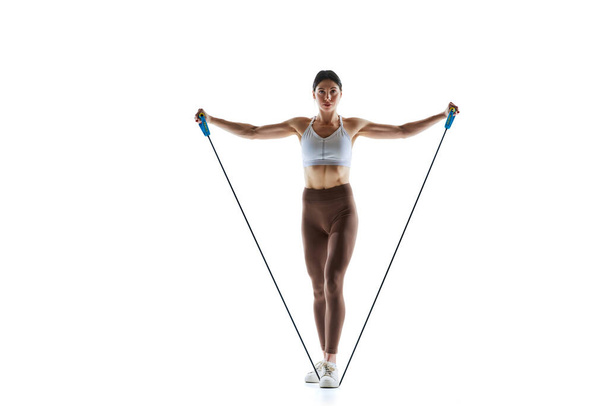 Female athlete in sportswear using resistance band for training against white studio background. Concept of sport and recreation, movement, self care, action, energy. Ad - Photo, Image