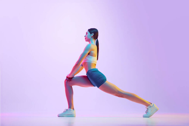 Young athlete woman doing lunges in motion in neon light against gradient studio background. Side view portrait. Concept of sport and recreation, movement, self care, action, energy. Ad - Φωτογραφία, εικόνα