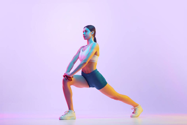 Young athlete woman doing lunges in motion in neon light against gradient studio background. Side view portrait. Concept of sport and recreation, movement, self care, action, energy. Ad - Photo, Image