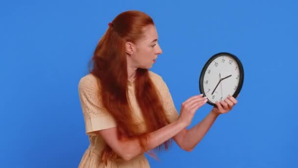 Young adult woman with anxiety checking time on clock, running late to work, being in delay, deadline. Redhead girl looking at hour, minutes, worrying to be punctual isolated on blue studio background - Footage, Video