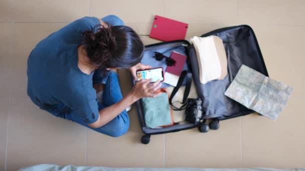 Vista superior del viajero femenino usando el teléfono inteligente mientras recoge cosas en la maleta antes del viaje - Imágenes, Vídeo