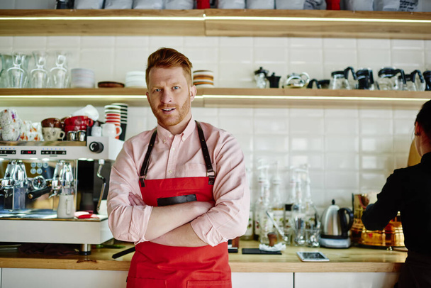 Mature redhead male in casual clothes and red apron standing in cafe kitchen with arms folded and looking at camera with smile - Fotó, kép
