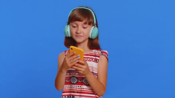 Young school girl listening music via headphones and dancing disco fooling around having fun expressive gesticulating hands. Preteen Caucasian child kid use smartphone app isolated on blue background - Footage, Video