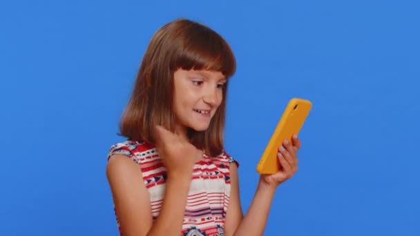 Nuori esiteini lapsi tyttö lapsi käyttää mobiili älypuhelin kirjoittamalla selailu sanoa Vau kyllä selville suuri iso voitto hyviä uutisia juhlia tekee voittaja ele. Pieni söpö lapset studio sininen tausta - Materiaali, video