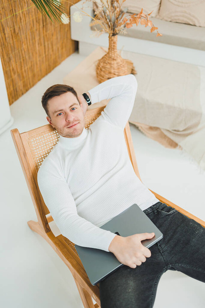 Mladý muž odpočívá doma, sedí v pohodlném křesle v obývacím pokoji a pracuje na notebooku, zatímco se dívá na svůj notebook. - Fotografie, Obrázek