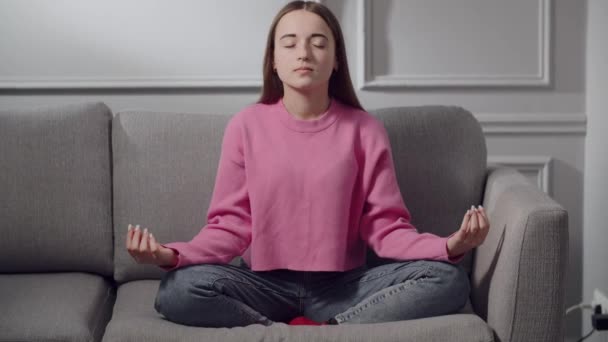 Ein süßes, konzentriertes Teenager-Mädchen praktiziert Yoga zu Hause. Eine junge Studentin sitzt in der Lotusstellung padmasana auf dem Sofa im Zimmer, atmet tief ein und schließt die Augen. Das Konzept des Nützlichen - Filmmaterial, Video