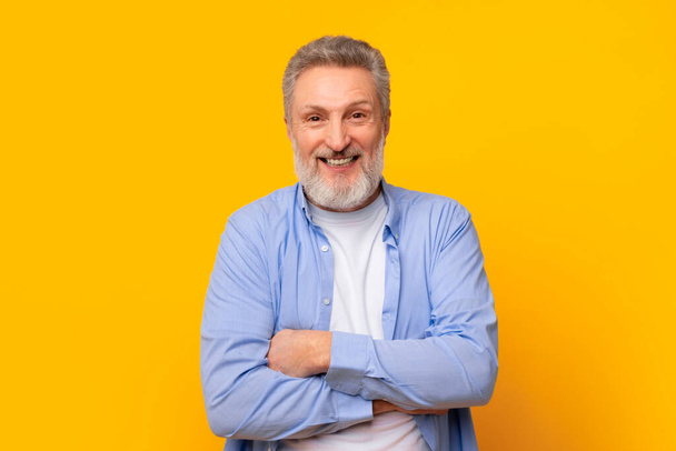 Успішна старша особа. Портрет щасливого зрілого європейця з сірою бородою, що стоїть з схрещеними руками, виражаючи впевненість і позитивність над жовтою стіною студії, посміхаючись камері - Фото, зображення