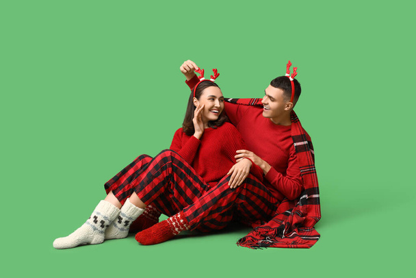 Happy young couple in Christmas pajamas and with plaid sitting against green background - Photo, Image