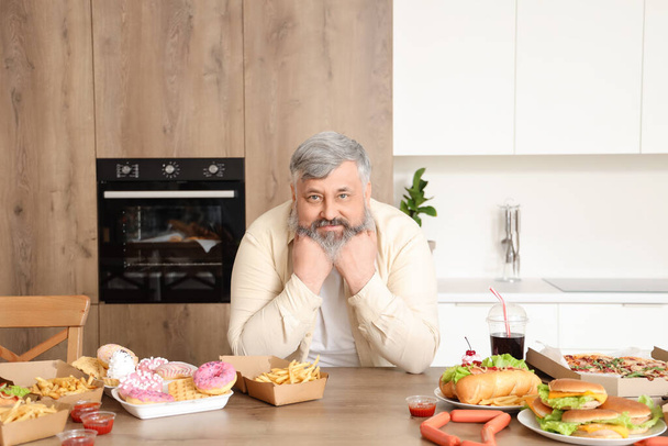 Избыточный взрослый мужчина за столом, полным нездоровой пищи на кухне. Концепция переедания - Фото, изображение