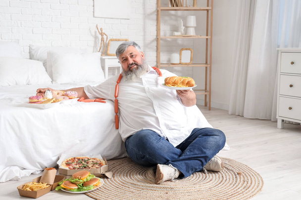 Dospělý muž s nadváhou a nezdravým jídlem v ložnici. Koncept přejídání - Fotografie, Obrázek