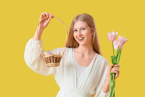 黄色の背景にスパアクセサリーとチューリップの花とイースターのバスケットを保持する美しい若い女性 - 写真・画像