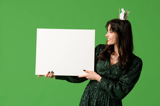 Hermosa mujer joven en vestido de fiesta elegante y corona de papel con tarjeta en blanco sobre fondo verde - Foto, Imagen