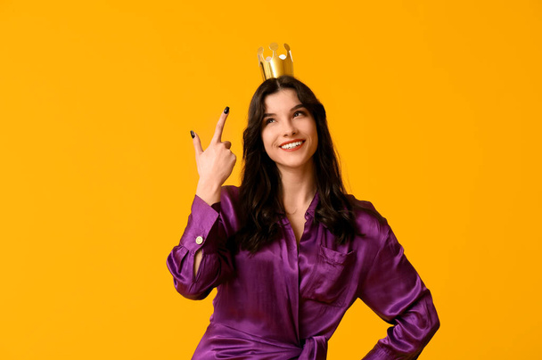 Beautiful young woman in stylish prom dress pointing at paper crown on yellow background - Photo, Image