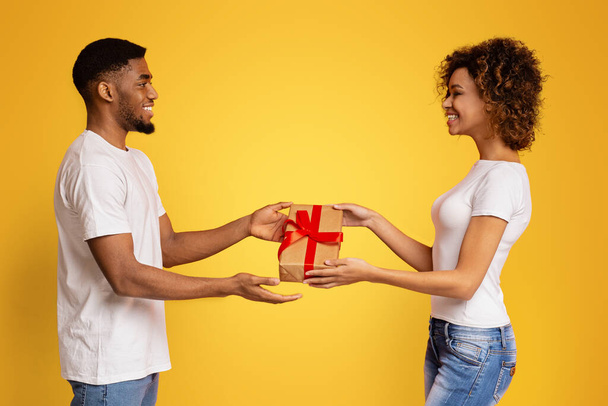 Happy black couple giving holiday gift to each other, orange studio background, free space - Photo, Image