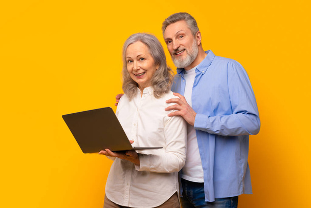 Χαμογελώντας ηλικιωμένοι σύζυγοι που στέκονται με τον υπολογιστή, ασχολούνται με την ηλεκτρονική μάθηση και την βιντεοκλήση στο laptop τους, κύλιση των μέσων κοινωνικής δικτύωσης και websurfing μαζί σε κίτρινο φόντο στούντιο - Φωτογραφία, εικόνα