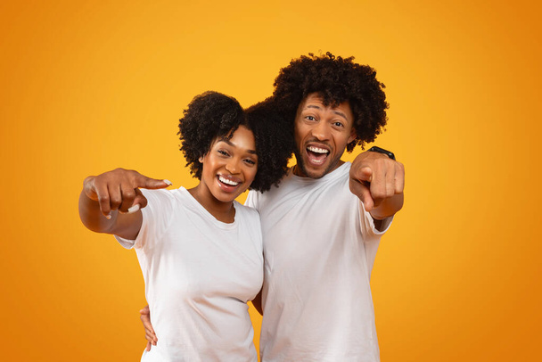Zabawna afrykańska para wskazująca na kamerę i śmiejąca się na żółtym tle studia. Szczęśliwy pewny siebie millenium czarny mężczyzna i kobieta obejmując i gestykulując - Zdjęcie, obraz