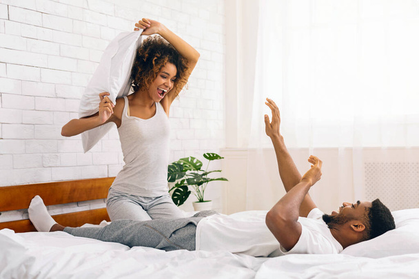 Pelea de almohadas alegre y divertida escena con una joven pareja afroamericana sonriente en una cama en casa - Foto, Imagen
