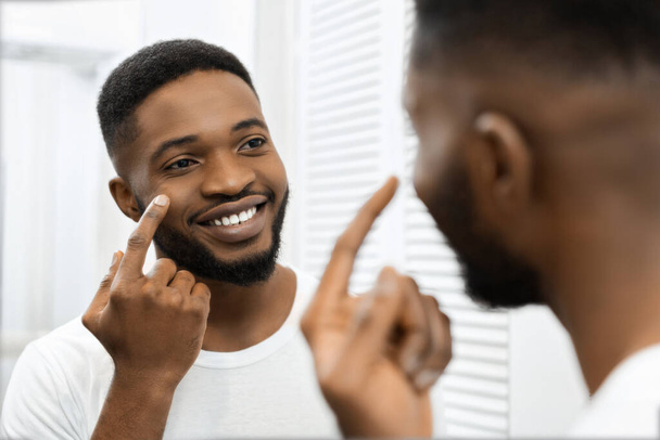 Ένας χαρούμενος Αφροαμερικάνος βάζει κρέμα προσώπου χαμογελώντας, αντανακλώντας ένα θετικό ξεκίνημα στην πρωινή του ρουτίνα. - Φωτογραφία, εικόνα