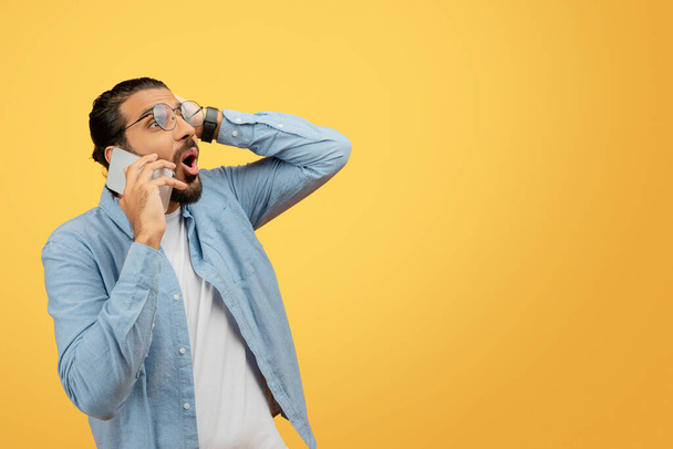 A perplexed young man in a casual denim shirt talking on phone and touching his head in confusion or forgetfulness against a bright yellow background - Photo, Image