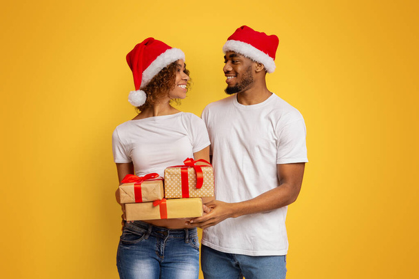 クリスマス休暇の準備ができて幸せなカップル。アフリカ系アメリカ人の男性と女性が多くのプレゼントを持って立ち、お互いに笑顔で、オレンジの背景 - 写真・画像