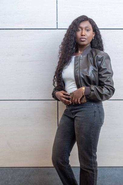 Una mujer africana equilibrada modela con confianza contra un muro de baldosas contemporáneo. Su chaqueta de cuero y jeans negros son el epítome de la elegancia urbana. Su cabello ondulado cae en cascada por sus hombros, completando el - Foto, imagen