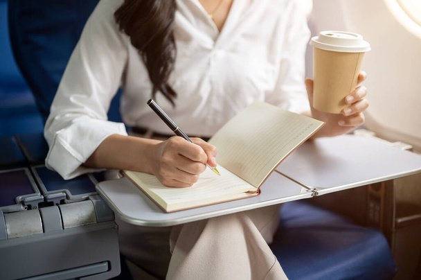 Una foto recortada de una mujer de negocios trabajando, tomando notas en su cuaderno, y bebiendo café mientras volaba a otra ciudad para un viaje de negocios. empresarios y conceptos de transporte - Foto, Imagen