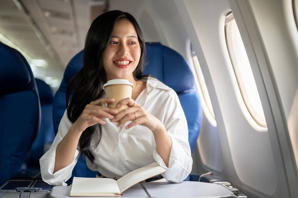 Atrakcyjna, elegancka azjatycka bizneswoman sączy kawę patrząc na widok za oknem samolotu, podróżując w podróż służbową. biznes, transport, luksusowy styl życia - Zdjęcie, obraz