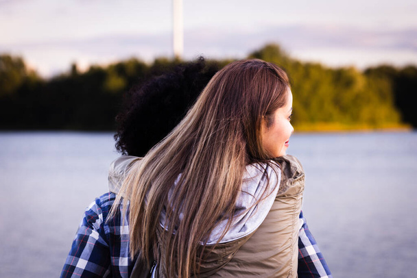 この画像は,穏やかな湖を眺めながら思考で失われた2人の友人の背中を描いています. 焦点は長い髪の女性にあり,カーリー髪の男性は後ろから見ることができます. - 写真・画像