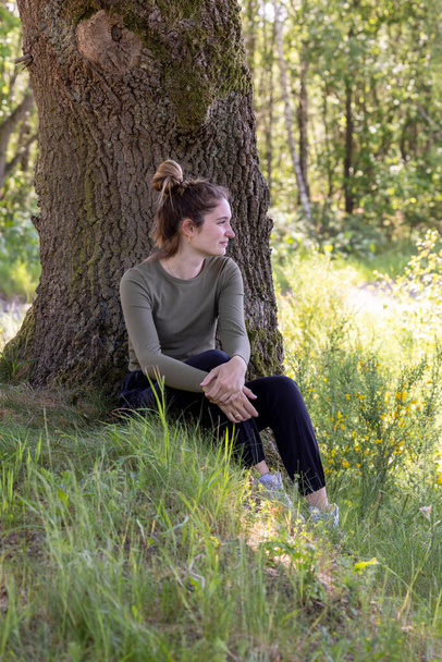 Dieses Bild zeigt eine kontemplative Frau, die am Fuß eines großen, strukturierten Baumes in einer grünen Waldkulisse sitzt. Ihr Blick richtet sich in die Ferne und suggeriert Introspektion oder tiefe Einblicke. - Foto, Bild