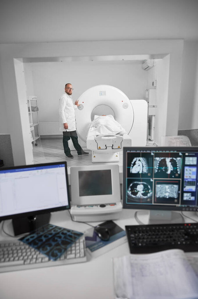 Orvosi komputertomográfia vagy MRI szkenner. Az orvos MRI-t készít, a beteg a kanapén fekszik. Férfi szakember fehér köntösben és szemüvegben, kamerába néz, mosolyog. A modern diagnosztika fogalma. - Fotó, kép