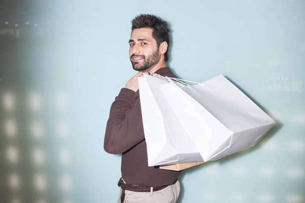 Minimal waist up portrait of smiling bearded man holding shopping bags standing against light blue background, camera flash - Photo, Image