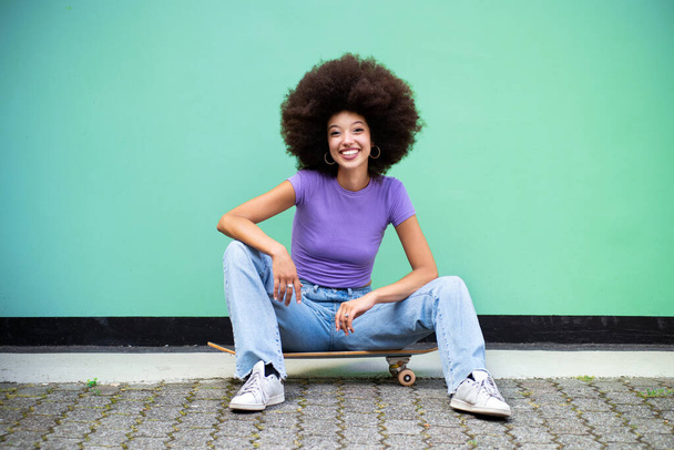Full body of cheerful young Moroccan female with Afro hairstyle and in casual clothes looking at camera while sitting in daylight on skateboard on street against blue wall - Foto, afbeelding