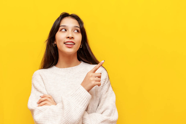 young asian girl with braces points with her hands to the side on a yellow isolated background, korean girl showing and advertising copy space - Zdjęcie, obraz