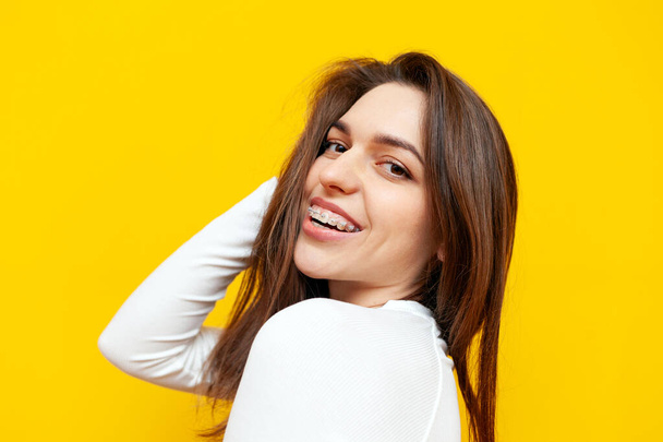 young woman with braces smiling on yellow isolated background, cheerful girl holding hand on hair and looking at camera - Photo, Image