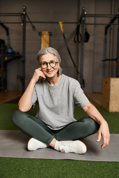 счастливая зрелая женщина в удобной спортивной одежде сидит со скрещенными ногами в спортзале и улыбается в камеру - Фото, изображение