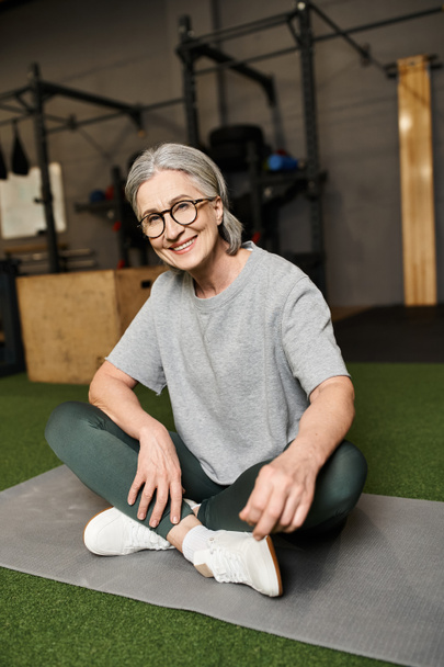 radosna dojrzała kobieta w okularach siedząca ze skrzyżowanymi nogami na podłodze w siłowni i uśmiechnięta do kamery - Zdjęcie, obraz