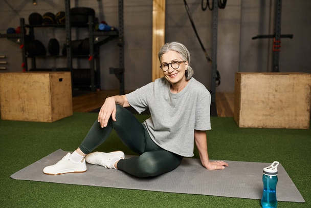 positive schöne reife Frau in Sportbekleidung mit Brille auf dem Boden sitzend und lächelnd in die Kamera - Foto, Bild