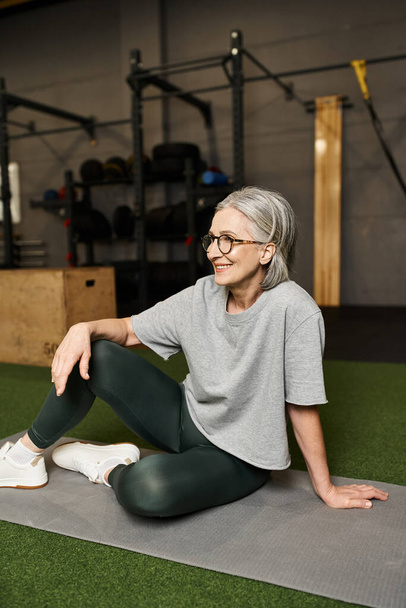 attractive merry mature woman in gray t shirt with glasses sitting on floor and looking away - Photo, Image