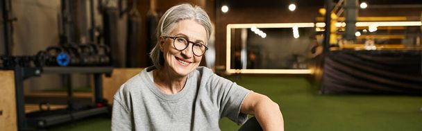 positive mature woman with glasses and gray hair sitting on floor and smiling at camera, banner - Photo, Image
