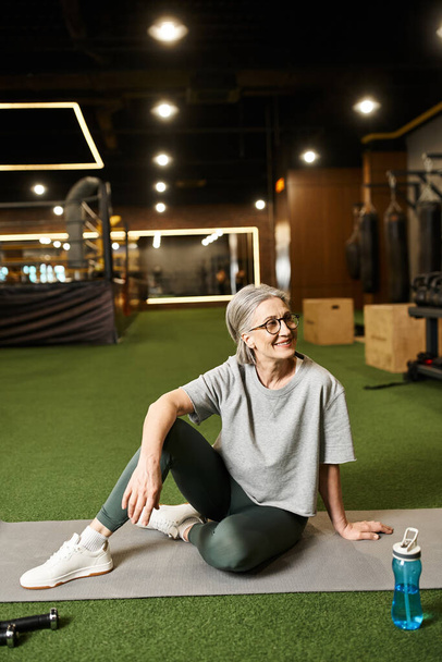 atractiva mujer madura feliz con el pelo gris y gafas sentado en el suelo en el gimnasio y mirando hacia otro lado - Foto, imagen