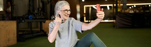 jolly mature sportswoman in gray t shirt with glasses taking selfies while holding dumbbells, banner - Photo, Image