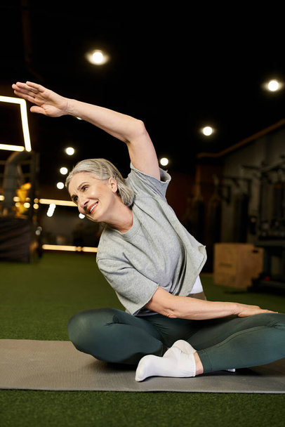 весёлая привлекательная пожилая женщина в удобной спортивной одежде с седыми волосами, растягивающими мышцы в спортзале - Фото, изображение