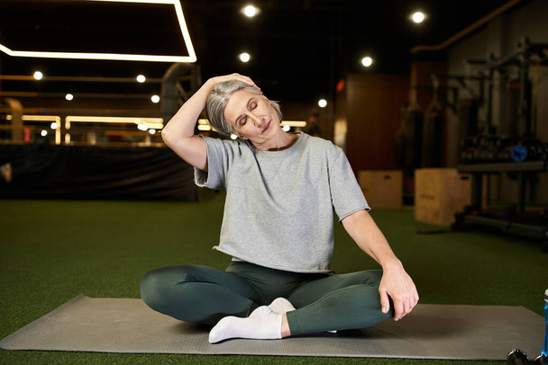 atlética atractiva mujer mayor en ropa deportiva estirando sus músculos mientras está sentada en el suelo en el gimnasio - Foto, imagen