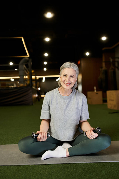 mujer madura alegre con el pelo gris en ropa deportiva haciendo ejercicio con pesas y mirando a la cámara - Foto, imagen