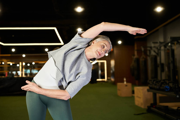 красивая, веселая, зрелая спортсменка в уютном наряде, растягивающая мышцы во время тренировки в спортзале - Фото, изображение