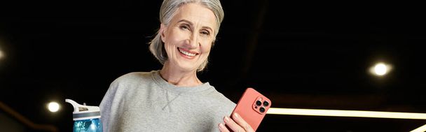 mujer madura alegre con pelo gris sosteniendo botella de agua y teléfono y sonriendo a la cámara, pancarta - Foto, Imagen